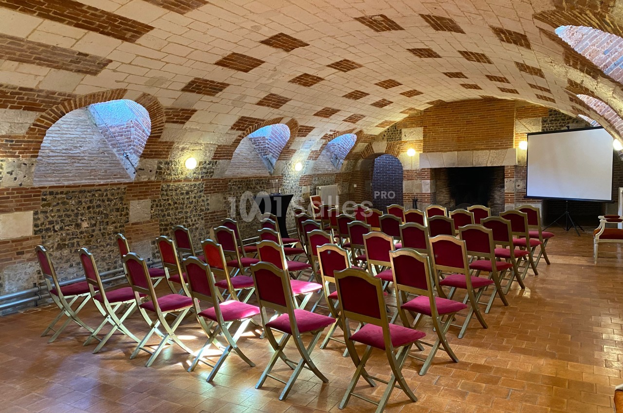 Salle voûtée en briques avec des rangées de chaises rouges disposées face à un écran de projection.