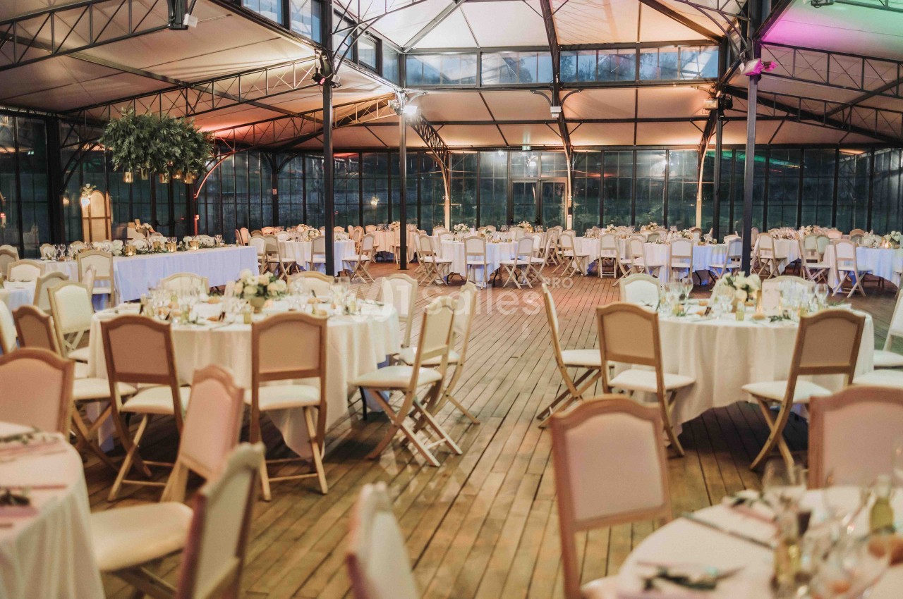 Salle de réception spacieuse avec tables rondes décorées de fleurs, chaises alignées et plafond en verrière.
