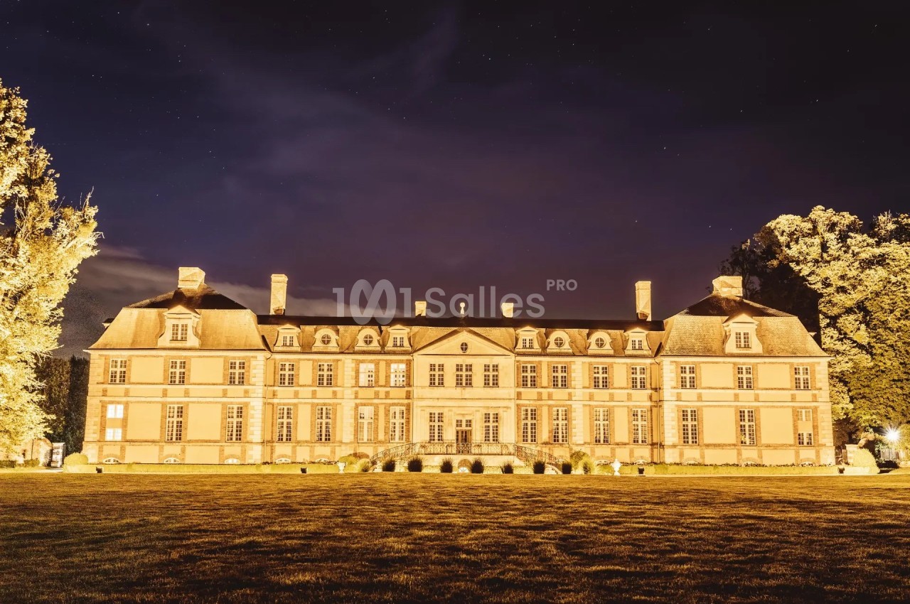 Façade éclairée d'un château en pierre entouré d'arbres, sous un ciel nocturne étoilé.