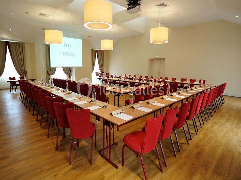 Salle de conférence lumineuse avec des tables disposées en U, des chaises rouges et un écran de projection.