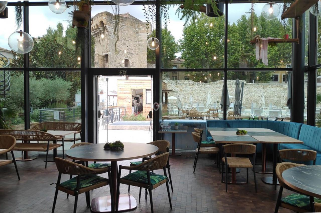 Location salle Aix-en-Provence (Bouches-du-Rhône) - Hôtel Aquabella & Spa #13 Intérieur d'un café avec des tables en bois, grandes baies vitrées et vue sur une terrasse et des ruines historiques.