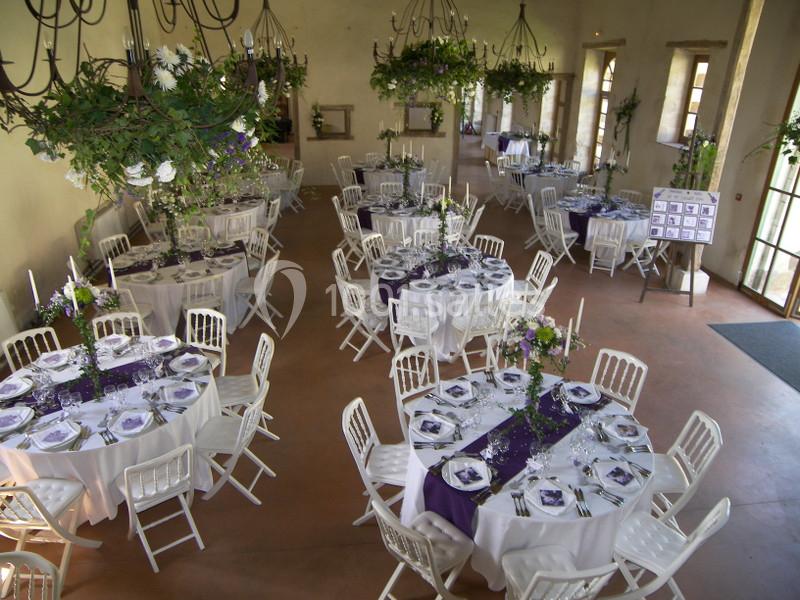 Salle de réception décorée avec des tables rondes, nappes blanches, chemins de table violets et chandeliers suspendus.