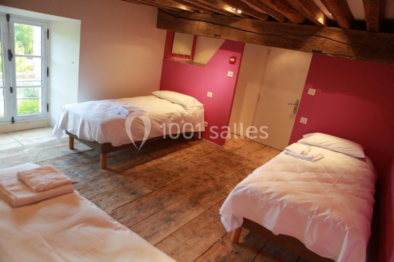 Chambre avec trois lits simples, parquet en bois, murs roses et blancs, et fenêtre donnant sur un espace extérieur verdoyant.