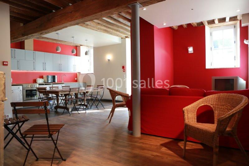 Salle lumineuse avec cuisine ouverte, murs rouges, poutres apparentes, mobilier en bois et fauteuils en osier.