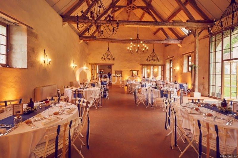 Salle de réception lumineuse avec tables décorées, chaises blanches et poutres apparentes sous un plafond en bois.