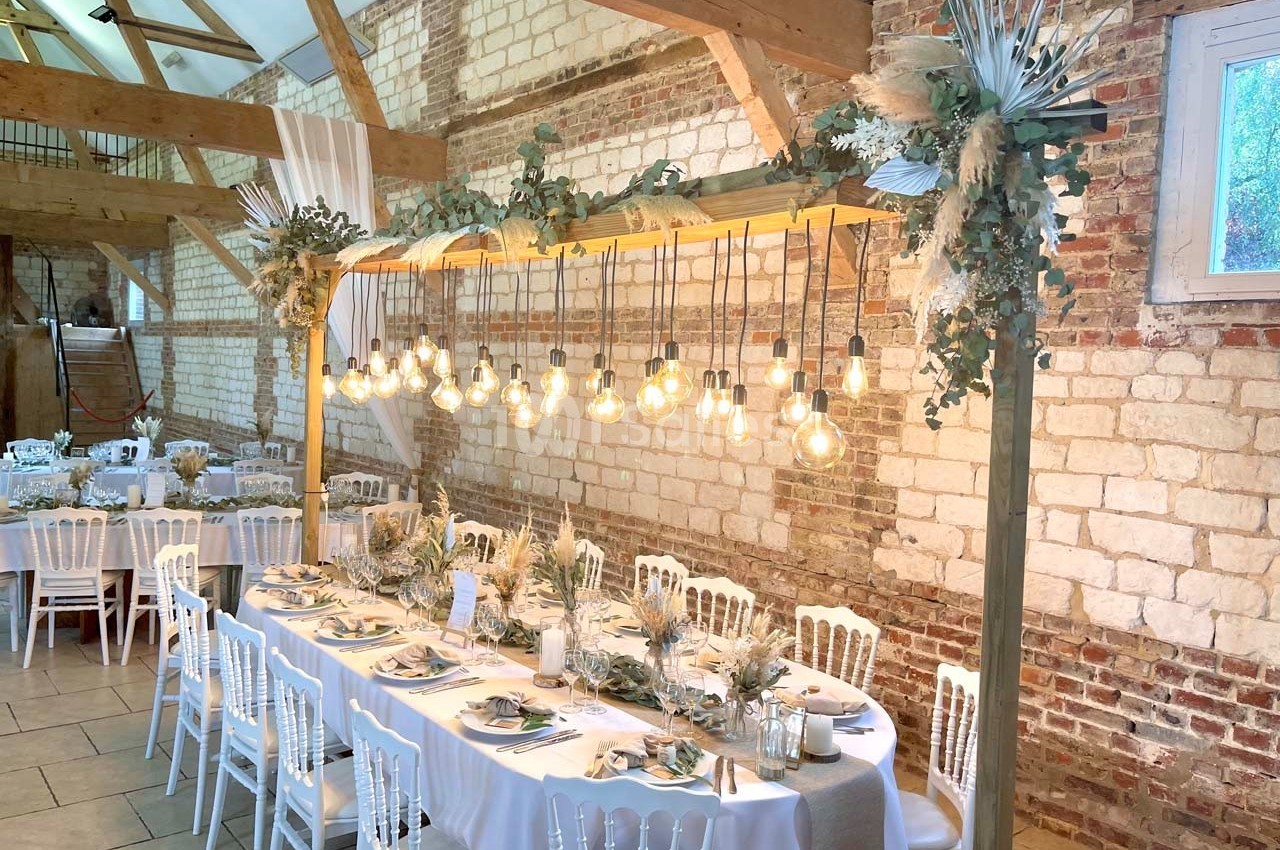 Table décorée pour un repas festif dans une salle en briques avec guirlande d'ampoules suspendues et fleurs séchées.