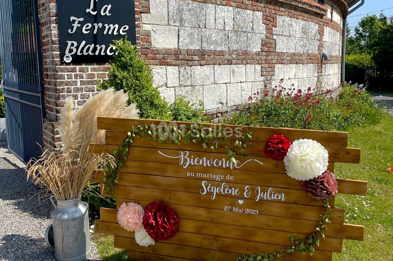Panneau en bois décoré de fleurs et feuillages, annonçant un mariage devant un mur en briques et l'enseigne ’La Ferme…