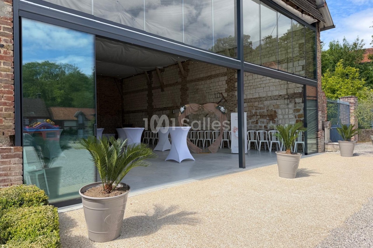 Salle de réception ouverte avec murs en pierre, tables hautes blanches et plantes en pot sur une terrasse en gravier.