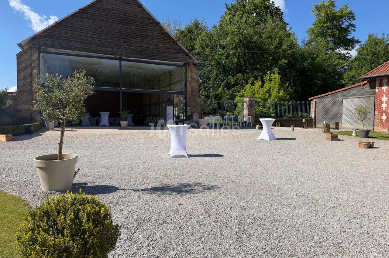 Cour extérieure en gravier avec une grange ouverte, des tables hautes blanches et des plantes en pot, entourée de verdure.