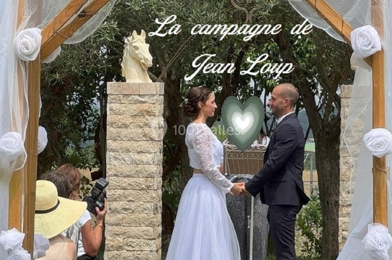 Un couple échange ses vœux sous une arche décorée de voilages et de fleurs, en extérieur, devant des invités.