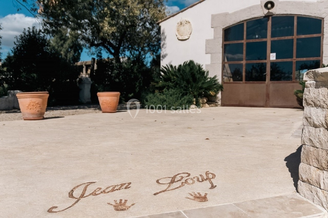 Entrée d'un bâtiment en pierre avec une terrasse gravée du texte ’Jean Loup’ et des pots de fleurs en terre cuite.