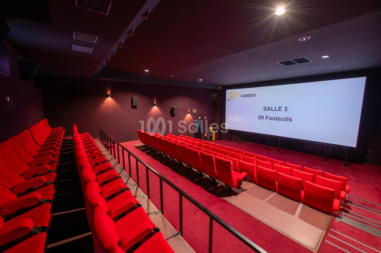 Salle de cinéma vide avec des sièges rouges, écran blanc affichant ’Salle 3, 89 fauteuils’ et éclairage tamisé.