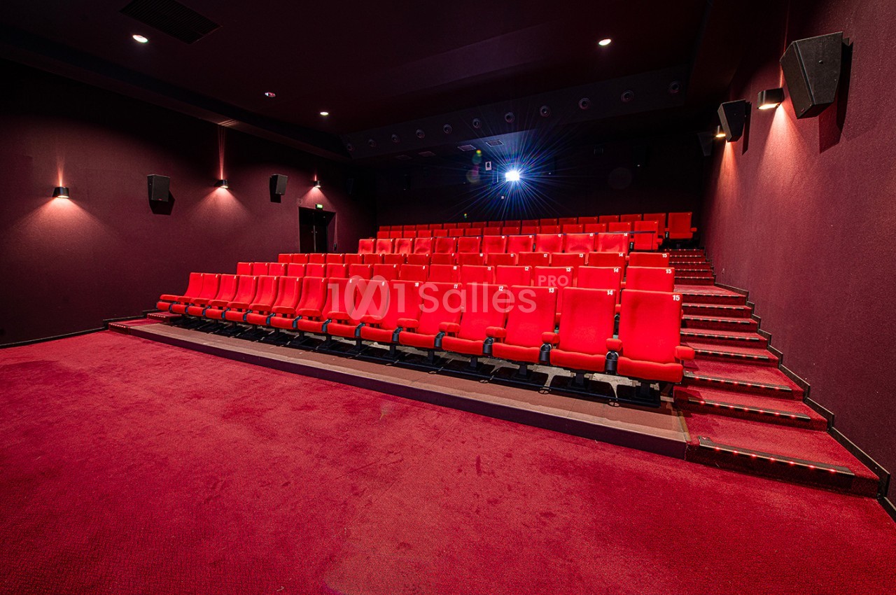 Salle de cinéma vide avec des rangées de sièges rouges et un projecteur allumé au fond.