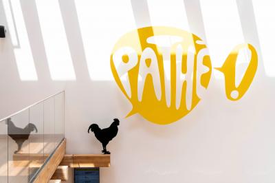 Miniature Location salle Paris 13 (Paris) - Pathé Les Fauvettes #12 Silhouette d'un coq noir sur une rambarde en bois devant un mur blanc avec le logo jaune ’Pathé !’ éclairé par la lumière.
