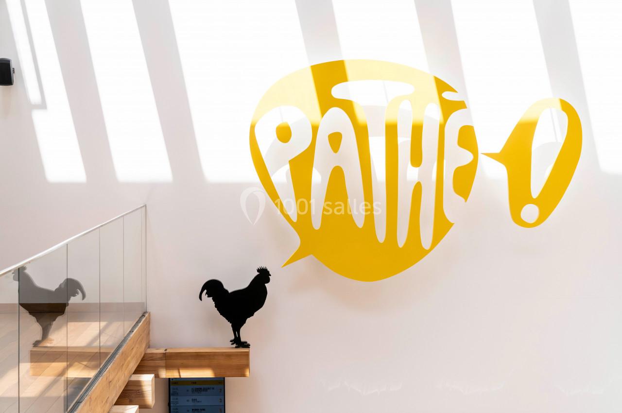 Location salle Paris 13 (Paris) - Pathé Les Fauvettes #12 Silhouette d'un coq noir sur une rambarde en bois devant un mur blanc avec le logo jaune ’Pathé !’ éclairé par la lumière.