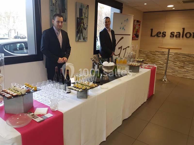 Deux serveurs devant une table dressée avec boissons, verrines et couverts dans une salle lumineuse.