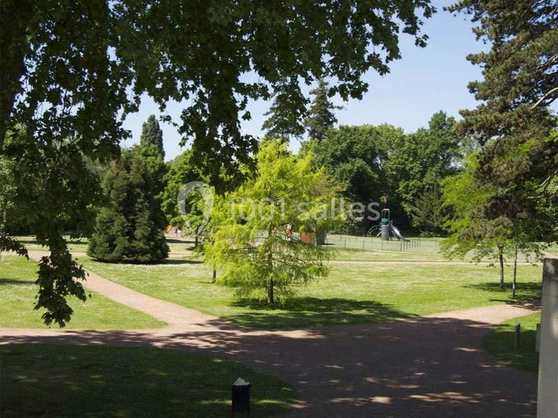 Vue d'un parc avec des arbres, des allées en terre et une aire de jeux visible en arrière-plan.