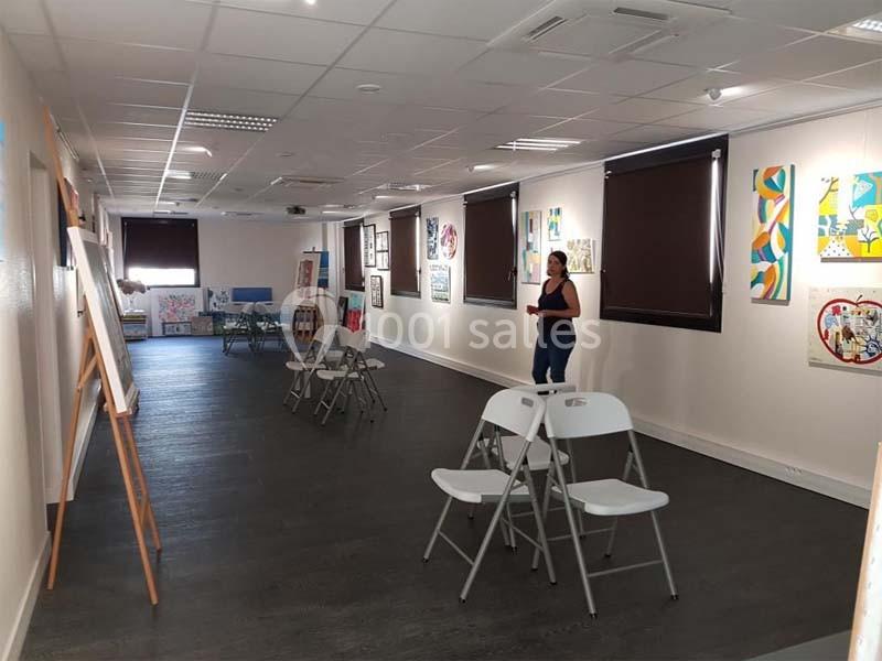 Salle d'exposition avec des tableaux accrochés aux murs, des chaises pliantes et une personne debout.