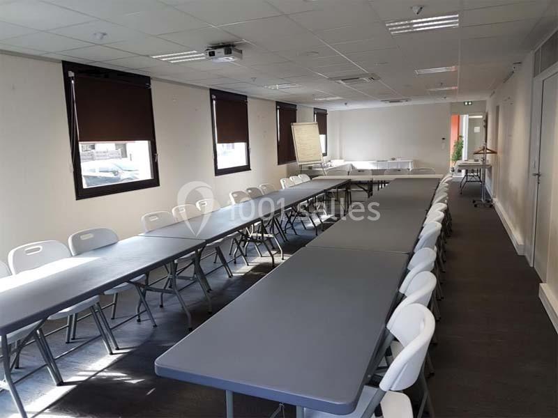 Salle de réunion lumineuse avec de longues tables grises alignées, entourées de chaises blanches pliantes.