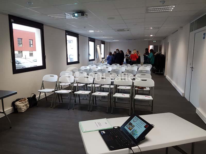 Salle de réunion avec des chaises alignées, un ordinateur portable ouvert et un groupe de personnes en discussion au fond.