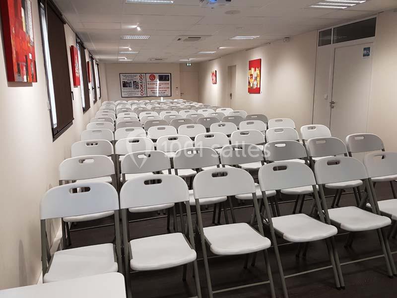 Salle de conférence avec des rangées de chaises blanches alignées face à un écran et une porte au fond.