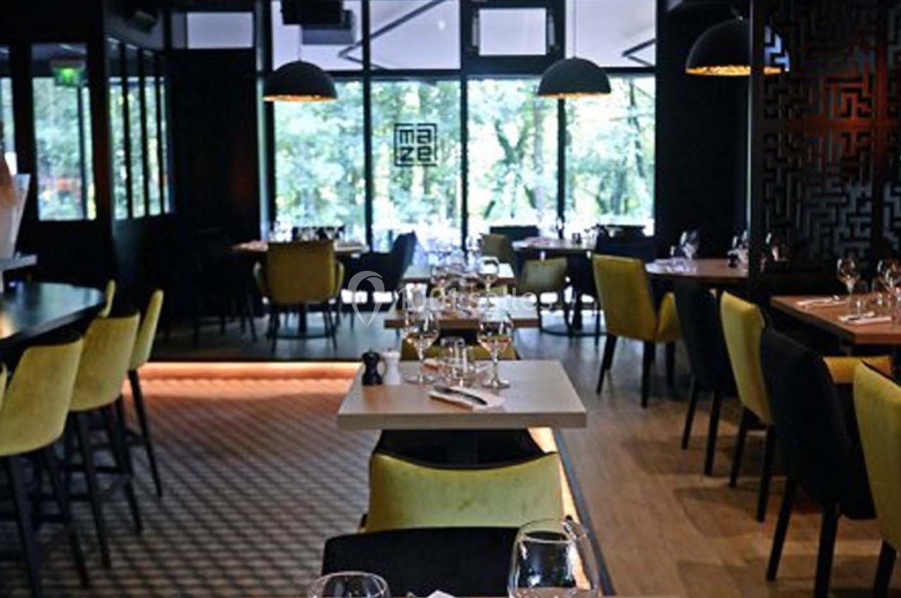 Salle de restaurant moderne avec tables dressées, chaises jaunes et noires, éclairage tamisé et grandes baies vitrées.