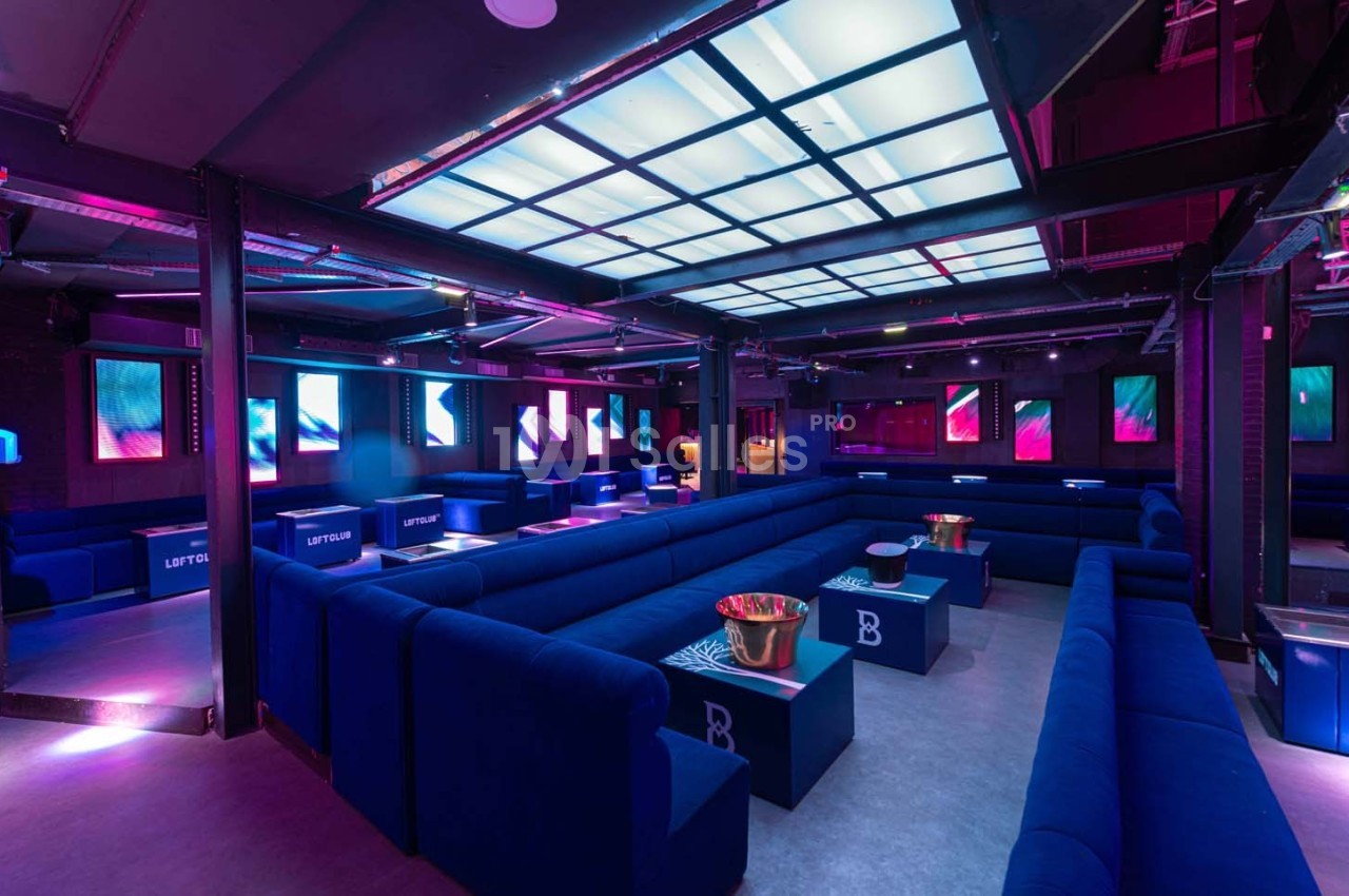 Salle de club moderne avec canapés bleus, tables basses et éclairage coloré diffusé par des écrans et des plafonniers.