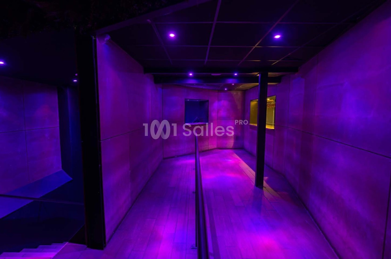 Couloir sombre éclairé par des lumières violettes, avec un sol en bois et des murs lisses.