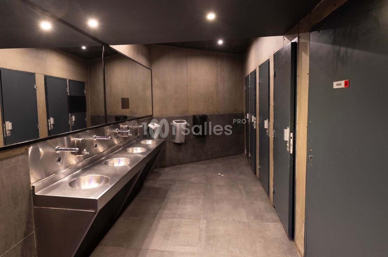 Salle de toilettes modernes avec lavabos en acier inoxydable, miroirs, cabines fermées et éclairage au plafond.
