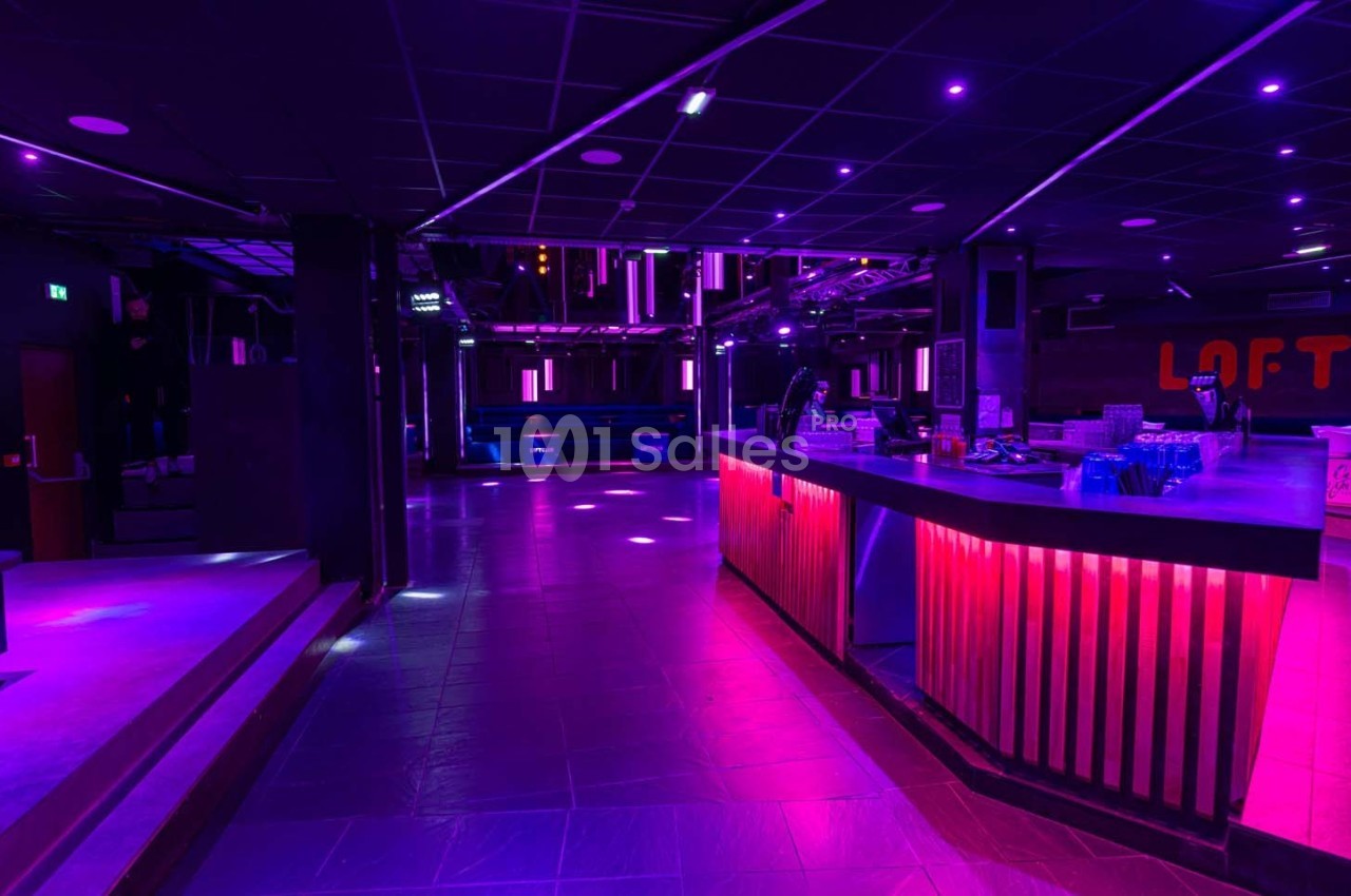 Salle de discothèque vide avec éclairage violet et rose, bar illuminé et piste de danse en arrière-plan.