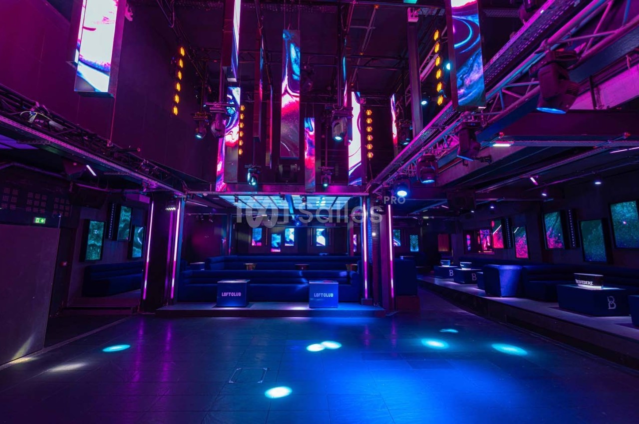Salle de discothèque vide avec éclairage néon coloré, écrans lumineux et banquettes alignées le long des murs.