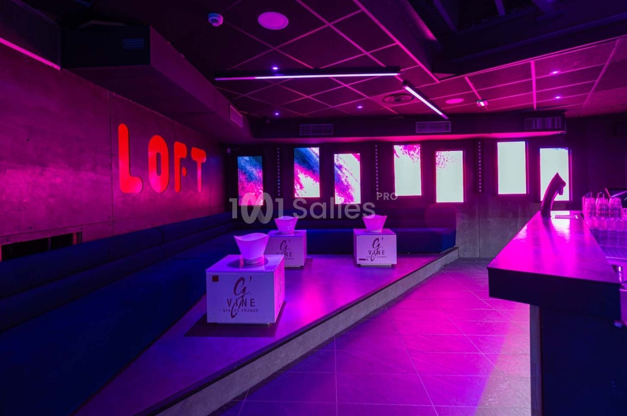 Salle de club avec éclairage rose, banquettes bleues, tables basses et écrans lumineux au mur.