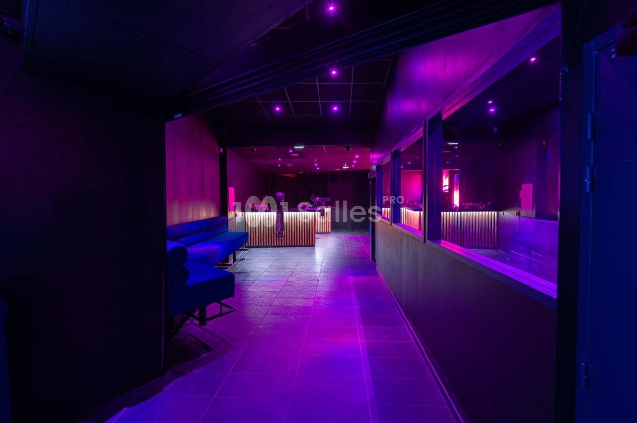 Salle sombre avec éclairage violet, comptoir lumineux en bois, banquettes bleues et murs noirs.