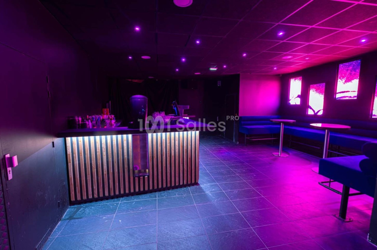 Salle sombre avec éclairage violet, bar en bois éclairé, tables et banquettes bleues le long des murs.