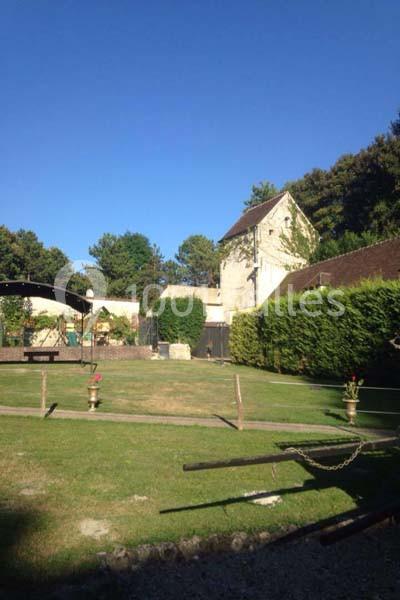 Cour verdoyante avec pelouse, arbres, bâtiments en pierre et pergola sous un ciel bleu dégagé.