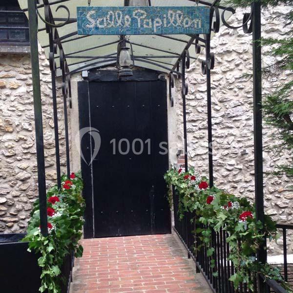 Passage couvert en fer forgé menant à une porte noire, bordé de fleurs rouges et de feuillage.