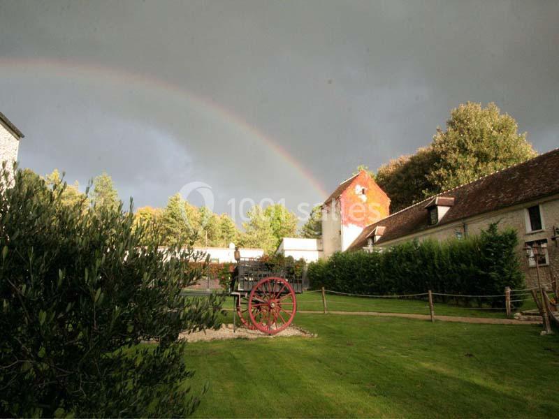 Arc-en-ciel au-dessus d'un jardin verdoyant avec une charrette ancienne et des bâtiments en pierre en arrière-plan.