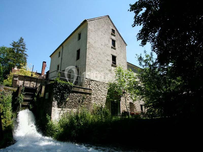 Bâtiment en pierre de plusieurs étages près d'une chute d'eau, entouré de végétation sous un ciel bleu.