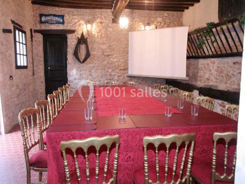 Salle de réunion rustique avec table rectangulaire couverte d'une nappe rouge, chaises dorées et écran de projection.