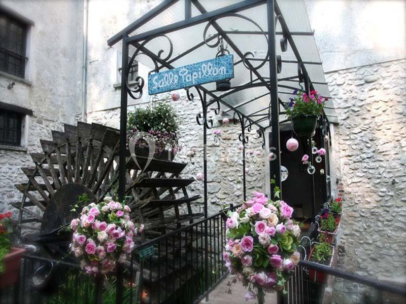 Passage couvert décoré de fleurs roses avec une roue à aubes en bois et un mur en pierre blanche en arrière-plan.