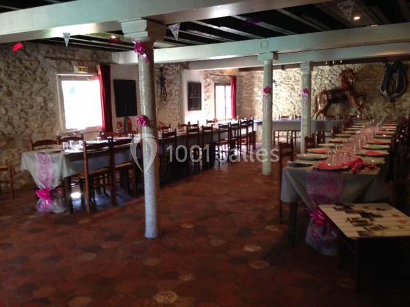 Salle de réception avec tables alignées, nappes grises et décorations roses, murs en pierre apparente.