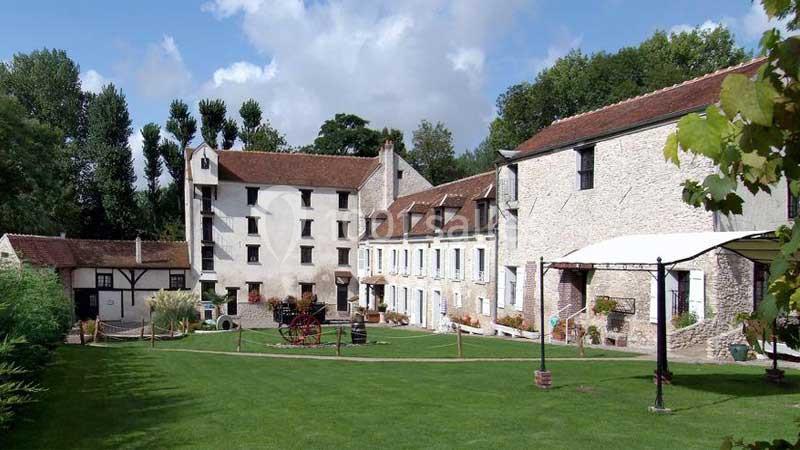 Bâtisse en pierre avec toits en tuiles entourée d'un jardin verdoyant et d'arbres sous un ciel partiellement nuageux.