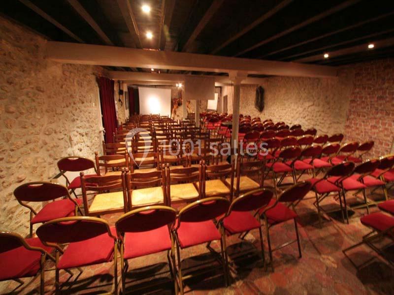 Salle avec des rangées de chaises rouges alignées, murs en pierre et plafond avec poutres apparentes.