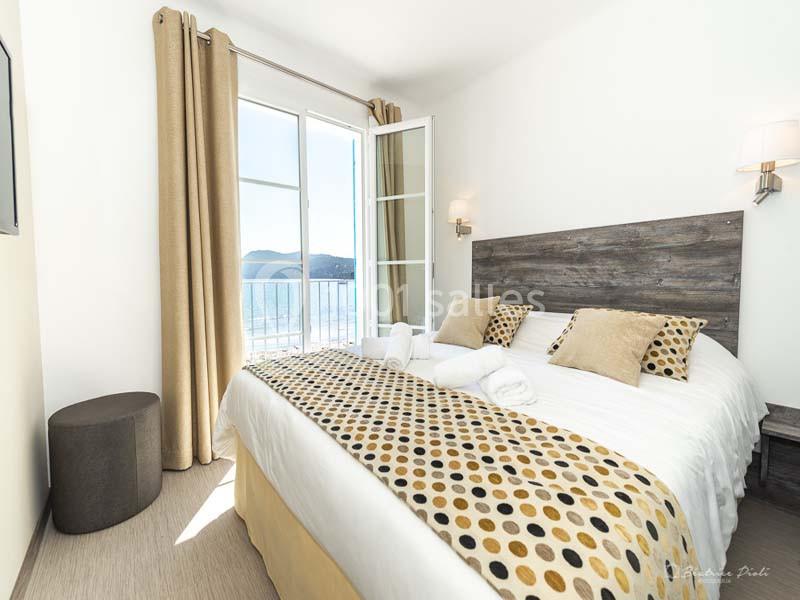 Chambre lumineuse avec lit double, décor beige et fenêtre ouverte offrant une vue sur la mer et les collines.