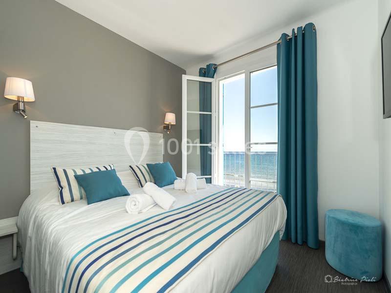 Chambre lumineuse avec lit double, décor bleu et blanc, fenêtres ouvertes offrant une vue sur la mer.