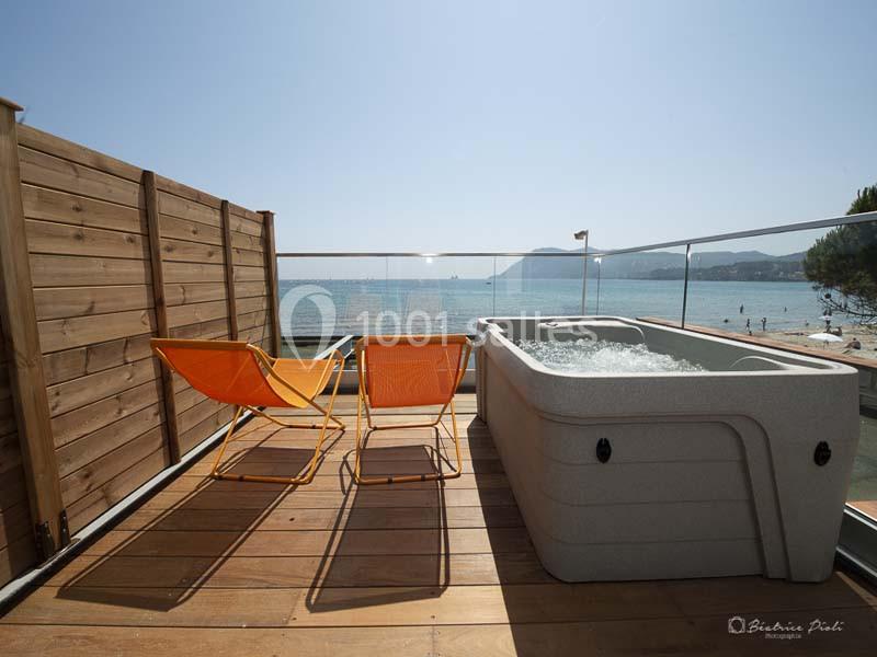 Terrasse en bois avec jacuzzi, deux chaises longues orange et vue sur la mer par une balustrade en verre.