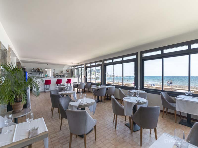 Salle de restaurant lumineuse avec vue sur la mer, tables dressées et bar en arrière-plan.
