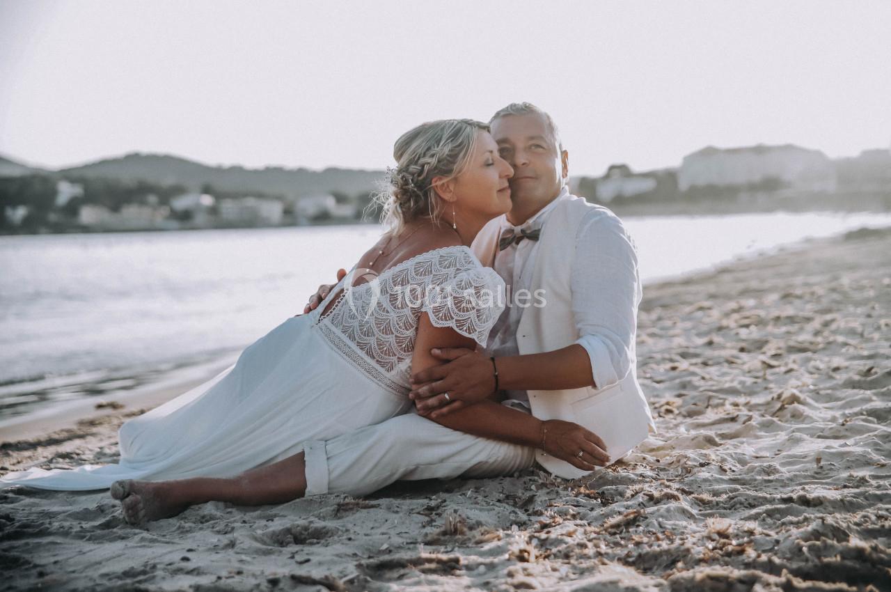 Un couple en tenue de mariage assis sur une plage au coucher du soleil, entouré d'un paysage côtier.