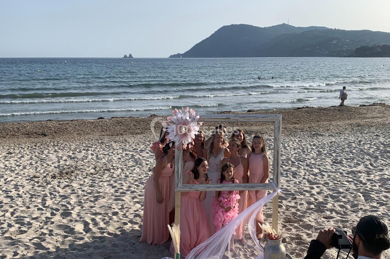 Groupe de personnes en robes roses posant derrière un cadre sur une plage avec la mer et des collines en arrière-plan.