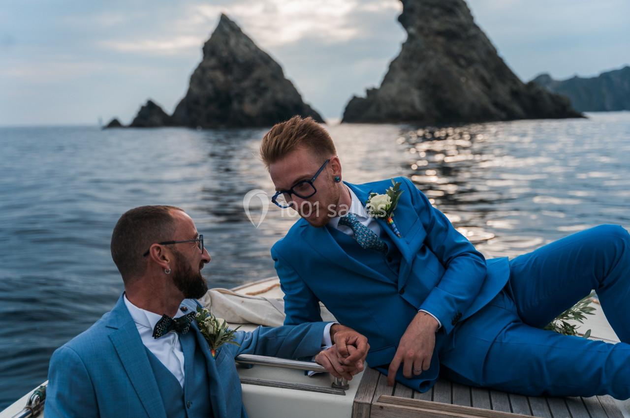 Deux hommes en costume bleu se tiennent la main sur un bateau, avec des rochers et la mer en arrière-plan.